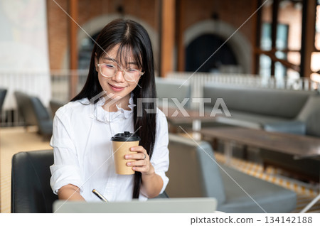 Asian woman student with eyeglasses holding pen and coffee while looking at laptop on table in cafe. Asian woman student with eyeglasses holding pen and coffee while looking at laptop on table in cafe. 134142188