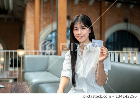 Pretty asian woman holding or showing a credit card while sitting on sofa in a cafe or coffee shop. Pretty asian woman holding or showing a credit card while sitting on sofa in a cafe or coffee shop. 134142250