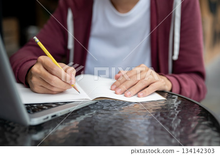 Close up of a man student holding pen writing in notebook over laptop on table in a cafe or library. Close up of a man student holding pen writing in notebook over laptop on table in a cafe or library. 134142303