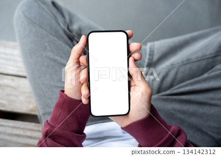 A male hands holding blank screen smartphone over folding legs while sitting outdoor on wooden bench A male hands holding blank screen smartphone over folding legs while sitting outdoor on wooden bench 134142312