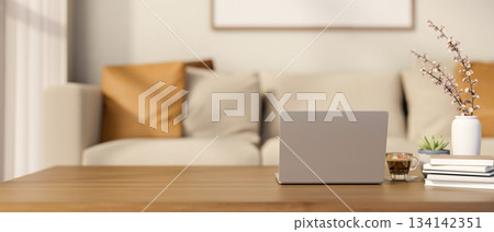 Laptop and a glass of coffee on wooden table with books and vase across sofa in blurred living room. 134142351
