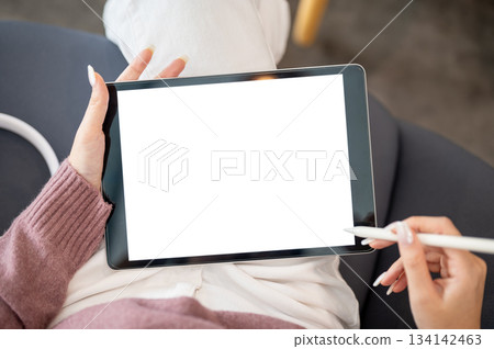 Close up of woman artist or student's hands holding tablet and stylus while sitting on sofa in cafe. Close up of woman artist or student's hands holding tablet and stylus while sitting on sofa in cafe. 134142463