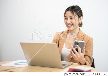 Asian businesswoman office worker holding a pen under chain looking at laptop on table in white room Asian businesswoman office worker holding a pen under chain looking at laptop on table in white room 134143137