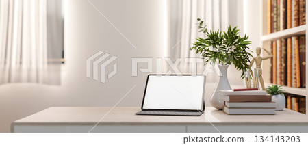 White screen tablet and books aside plant vase on wooden table aside shelf in a bright living room. 134143203