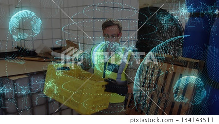 Wearing safety vest, worker holding yellow crate in warehouse, with pallets and holographic overlay 134143511