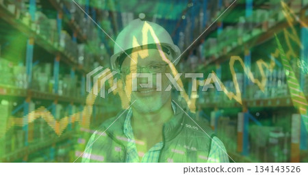 Standing warehouse manager wearing white helmet, glasses, gray vest in aisle with financial charts 134143526