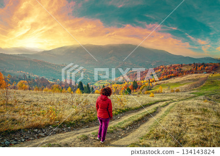 Mountain range in autumn. Young woman stands on dirt road with her back to the camera and looks at beautiful sunset over the mountains 134143824