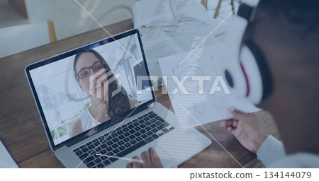 Colleague wearing headphones calling on laptop reviewing printed diagram at office desk, copy space 134144079