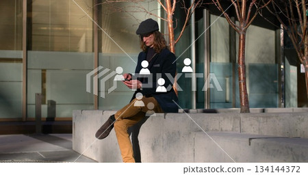 Browsing smartphone man with newsboy cap sitting on bench in plaza, among planters with bare trees Browsing smartphone man with newsboy cap sitting on bench in plaza, among planters with bare trees 134144372