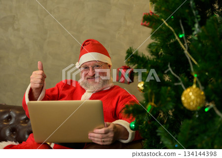 Santa claus giving thumbs up while using laptop computer. Man sitting on brown leather sofa next to festive decorated christmas tree in living room at home 134144398