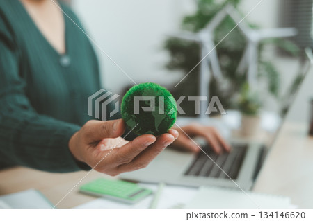 Eco-friendly office scene with person working on laptop near solar panel and wind turbine model, representing green business, renewable energy innovation and sustainable technology workspace. 134146620