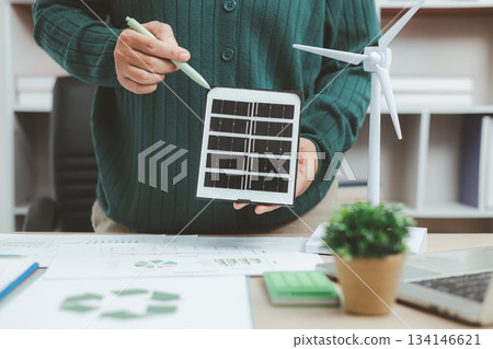 Eco-friendly office scene with person working on laptop near solar panel and wind turbine model, representing green business, renewable energy innovation and sustainable technology workspace. 134146621
