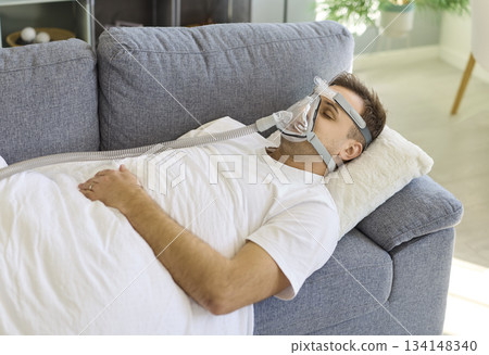 Man resting on sofa with CPAP oxygen mask for sleep apnea therapy at home Man resting on sofa with CPAP oxygen mask for sleep apnea therapy at home 134148340