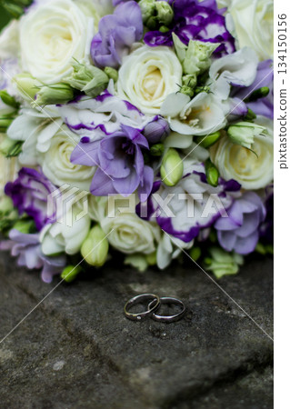 Wedding rings resting on stone with bridal bouquet in background Wedding rings resting on stone with bridal bouquet in background 134150156