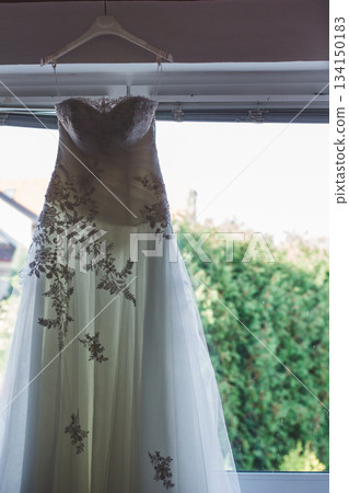 Elegant white wedding dress hanging near window, waiting for the bride Elegant white wedding dress hanging near window, waiting for the bride 134150183