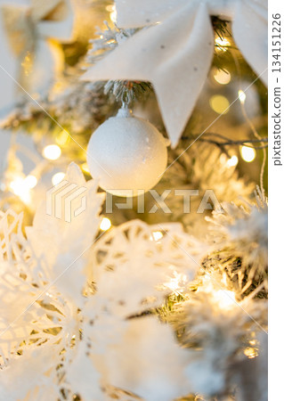 Snow-covered Christmas tree with white Christmas tree decorations close-up with warm twinkling lights. Decorations for the New Year and Christmas 134151226