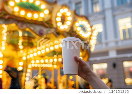 Woman hand holding disposable takeaway coffee cup with Christmas carousel on Christmas fair in the background. Mockup, space for text. Blurred background with Christmas lights. Recyclable packaging. 134151235