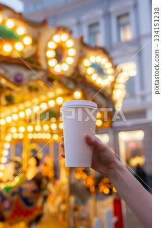 Woman hand holding disposable takeaway coffee cup with Christmas carousel on Christmas fair in the background. Mockup, space for text. Blurred background. Recyclable packaging Woman hand holding disposable takeaway coffee cup with Christmas carousel on Christmas fair in the background. Mockup, space for text. Blurred background. Recyclable packaging 134151238