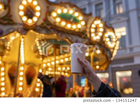 Woman hand holding disposable takeaway coffee cup with Christmas carousel on Christmas fair in the background. Mockup, space for text. Blurred background. Recyclable packaging. 134151247