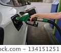 Close up of person refueling a car at a gas station using a green fuel nozzle. Concept of transportation, energy, travel readiness, routine, technology, and modern lifestyle. 134151251