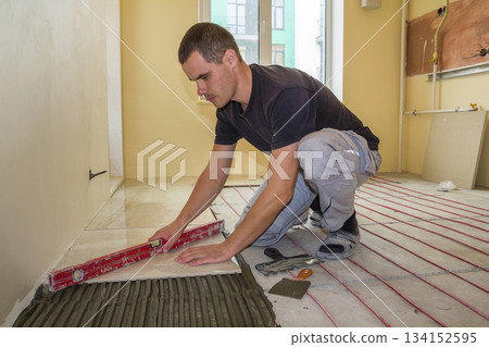 Young worker tiler installing ceramic tiles using lever on cement floor with heating red electrical cable wire system. Home improvement, renovation and construction, comfortable warm home concept. Young worker tiler installing ceramic tiles using lever on cement floor with heating red electrical cable wire system. Home improvement, renovation and construction, comfortable warm home concept. 134152595