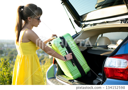 Young woman in summer dress putting green suitcase inside her car trunk. Travel and vacations concept. Young woman in summer dress putting green suitcase inside her car trunk. Travel and vacations concept. 134152598