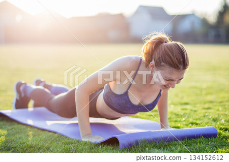 Young sportive woman in sports clothes training in field at sunrise. Girl standing in plank position with pain expression on grass in a city park. 134152612