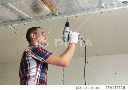 Young man in goggles fixing drywall suspended ceiling to metal frame using electrical screwdriver on ceiling insulated with shiny aluminum foil. Renovation, construction, do it yourself concept. Young man in goggles fixing drywall suspended ceiling to metal frame using electrical screwdriver on ceiling insulated with shiny aluminum foil. Renovation, construction, do it yourself concept. 134152626
