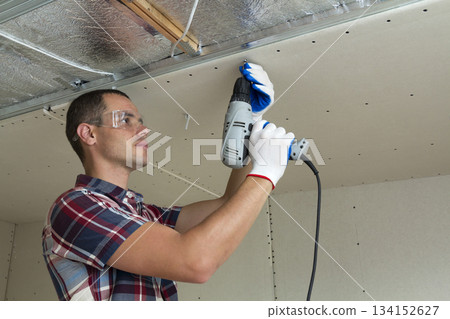 Young man in goggles fixing drywall suspended ceiling to metal frame using electrical screwdriver on ceiling insulated with shiny aluminum foil. Renovation, construction, do it yourself concept. Young man in goggles fixing drywall suspended ceiling to metal frame using electrical screwdriver on ceiling insulated with shiny aluminum foil. Renovation, construction, do it yourself concept. 134152627