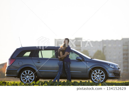 Young couple in love, slim attractive woman with long ponytail and handsome man standing together at silver car in green field on blurred apartment building and clear sky copy space background. 134152629