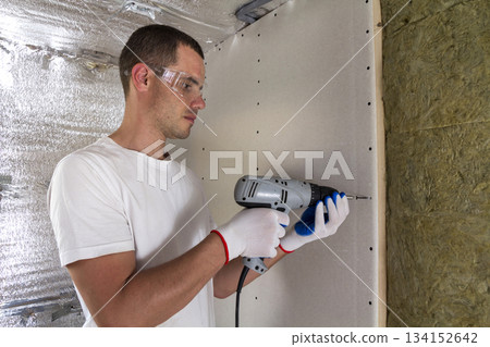 Worker in goggles with screwdriver working on insulation. Drywall on wall beams, insulating rock wool staff in wooden frame. Comfortable warm home, economy, construction and renovation concept. 134152642