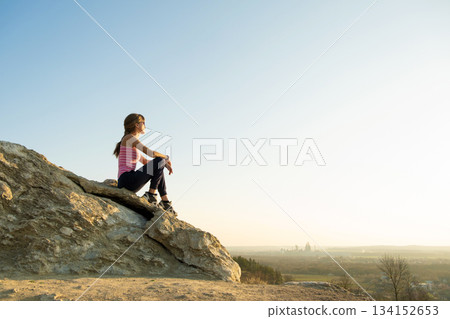 Woman hiker sitting on a steep big rock enjoying warm summer day. Young female climber resting during sports activity in nature. Active recreation in nature concept. Woman hiker sitting on a steep big rock enjoying warm summer day. Young female climber resting during sports activity in nature. Active recreation in nature concept. 134152653