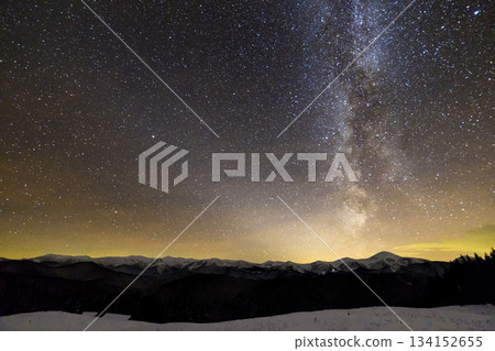 Winter mountains night landscape panorama. Milky Way bright constellation in dark starry sky, soft glow on horizon after sunset, magnificent mountain ridge snow-capped peaks, snowy steep woody hill. Winter mountains night landscape panorama. Milky Way bright constellation in dark starry sky, soft glow on horizon after sunset, magnificent mountain ridge snow-capped peaks, snowy steep woody hill. 134152655