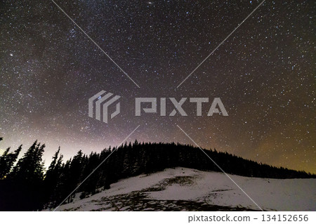 Winter mountains night landscape panorama. Milky Way bright constellation in dark blue starry sky over dark spruce pine trees forest, soft glow on horizon after sunset. Wide angle shot. Winter mountains night landscape panorama. Milky Way bright constellation in dark blue starry sky over dark spruce pine trees forest, soft glow on horizon after sunset. Wide angle shot. 134152656