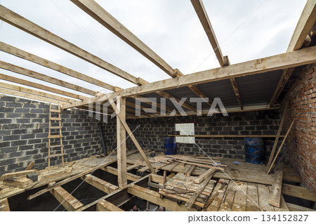 Two-storied building under construction and renovation. Perspective view of energy saving walls of large hollow foam insulation blocks and bricks, temporary wooden scaffolding, beams as roof frame. Two-storied building under construction and renovation. Perspective view of energy saving walls of large hollow foam insulation blocks and bricks, temporary wooden scaffolding, beams as roof frame. 134152827