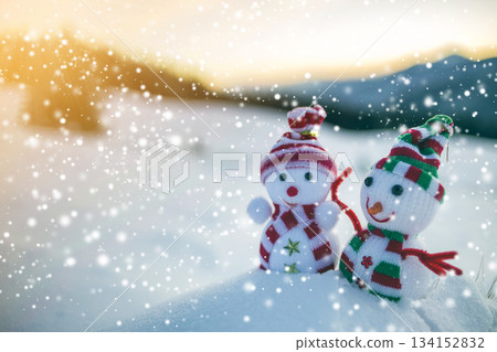 Two small funny toys baby snowman in knitted hats and scarves in deep snow outdoors on blurred mountains landscape background. Happy New Year and Merry Christmas greeting card theme. 134152832