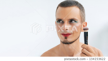 portrait of happy gay man with makeup and cosmetics on face. Handsome smiling transgender male with red lipstick on lips and bright eye shadows holds a blush brush on white background 134153622