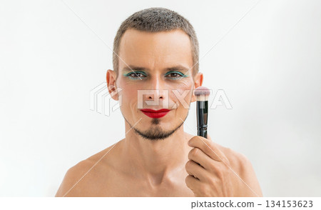 portrait of a feminine happy gay man with makeup and cosmetics on face. Handsome adult smiling transgender male with red lipstick on lips and bright eye shadows holds a blush brush on a white 134153623