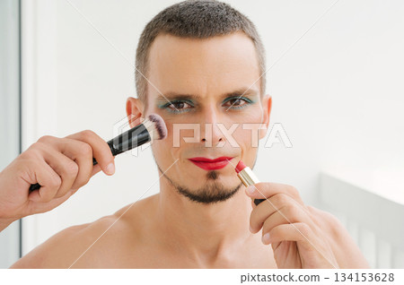 portrait of a happ gay man with makeup and cosmetics on face. A handsome young transgender male with bright eyeshadows paints lips with red lipstick and applies blush brush to face 134153628
