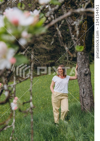 Smiling middle-aged woman standing near blooming tree in spring garden. Concept of female health, hormonal balance, wellness, and natural beauty in midlife 134154451
