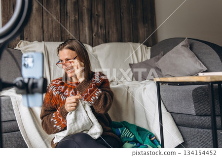 Middle aged woman talks online while knitting a cozy sweater at home. Family bond, digital communication, and peaceful hobby time blend into a warm  134154452