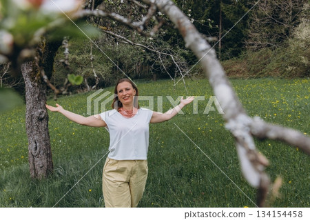 Smiling middle-aged woman standing near blooming tree in spring garden. Concept of female health, hormonal balance, wellness, and natural beauty in midlife 134154458