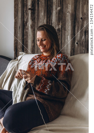 Woman sitting on a sofa and knitting with white yarn. Cozy handmade hobby, relaxation at home, quiet winter activity and creative selfcare concept Woman sitting on a sofa and knitting with white yarn. Cozy handmade hobby, relaxation at home, quiet winter activity and creative selfcare concept 134154472