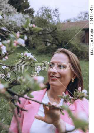 Smiling middle-aged woman standing near blooming tree in spring garden. Concept of female health, hormonal balance, wellness, and natural beauty in midlife Smiling middle-aged woman standing near blooming tree in spring garden. Concept of female health, hormonal balance, wellness, and natural beauty in midlife 134154473