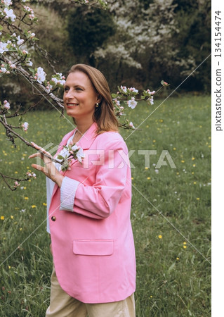 Smiling middle-aged woman standing near blooming tree in spring garden. Concept of female health, hormonal balance, wellness, and natural beauty in midlife Smiling middle-aged woman standing near blooming tree in spring garden. Concept of female health, hormonal balance, wellness, and natural beauty in midlife 134154474