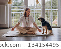 Middle aged woman practices Kundalini yoga at home with her dog nearby. Spiritual balance, feminine energy, harmony, mindfulness, and emotional wellness with animals 134154495