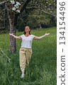 Smiling middle-aged woman standing near blooming tree in spring garden. Concept of female health, hormonal balance, wellness, and natural beauty in midlife 134154496