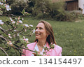 Smiling middle-aged woman standing near blooming tree in spring garden. Concept of female health, hormonal balance, wellness, and natural beauty in midlife 134154497