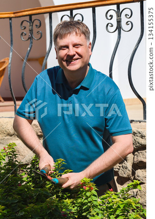 Adult Man Pruning Bushes in His Garden with Shears, Enjoying a Productive Day at Home Surrounded by Lush Greenery and Nature Adult Man Pruning Bushes in His Garden with Shears, Enjoying a Productive Day at Home Surrounded by Lush Greenery and Nature 134155375