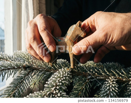 Strong hands gently place wooden horse toy on thick green branch highlighting the festive spirit of New Year in well lit room with sunlight. Strong hands gently place wooden horse toy on thick green branch highlighting the festive spirit of New Year in well lit room with sunlight. 134156979
